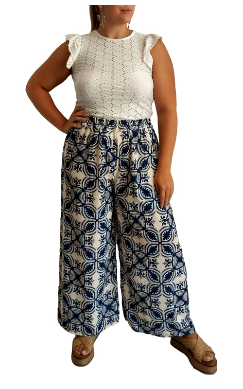Αυτό το κομψό σύνολο συνδυάζει αρμονικά την άνεση με το στυλ για τις καλοκαιρινές σας εμφανίσεις. Η παντελόνα με την φαρδιά γραμμή διαθέτει εντυπωσιακό μοτίβο σε λευκό και βαθύ μπλε, με γεωμετρικά σχέδια που θυμίζουν παραδοσιακά μεσογειακά μοτίβα. Η ψηλόμεση εφαρμογή κολακεύει τη σιλουέτα, ενώ το φαρδύ πόδι προσφέρει άνεση και αέρινη αίσθηση στις ζεστές μέρες. Συνδυάζεται τέλεια με το λευκό μπλουζάκι από δαντέλα με διάτρητο σχέδιο και χαριτωμένα βολάν στους ώμους, προσθέτοντας μια θηλυκή πινελιά στο σύνολο. Ιδανικό για καλοκαιρινές βόλτες, απογευματινούς καφέδες ή ακόμα και για χαλαρές βραδινές εξόδους στην παραλία. Μπορεί να συνδυαστεί με πλατφόρμες για πιο επίσημες περιστάσεις ή με επίπεδα σανδάλια για καθημερινές εμφανίσεις. Το φυσικό ύφασμα της παντελόνας εξασφαλίζει άνετη εφαρμογή και δροσερή αίσθηση, ενώ το έντονο μοτίβο της την καθιστά ξεχωριστό κομμάτι στη γκαρνταρόμπα σας.