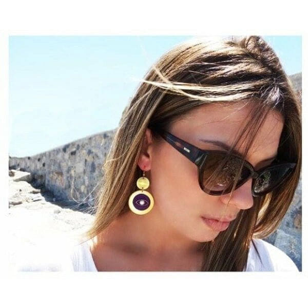 Σκουλαρίκια evil eye με μαργαριτάρια και ξύλο - 2996, Ανακαλύψτε τη μαγεία του evil eye με αυτά τα εντυπωσιακά χειροποίητα σκουλαρίκια που συνδυάζουν παραδοσιακή προστασία με μοντέρνο στυλ. Κάθε ζευγάρι σκουλαρικιών evil eye, σχεδιασμένο από την Ελληνίδα Σχεδιάστρια Κοσμημάτων Αικατερίνη Χαλκιαδάκη, αποτελεί μια μοναδική δημιουργία που ενώνει τη σύγχρονη αισθητική με την αρχαία ελληνική παράδοση.
Τα κρεμαστά σκουλαρίκια διαθέτουν δύο κομψούς κύκλους από ξύλο σε χρυσαφένια απόχρωση, με το κάτω τμήμα να φέρει εντυπωσιακό evil eye σε βαθύ μοβ χρώμα και λευκή πέρλα στο κέντρο. Η γεωμετρική σύνθεση και η αντίθεση των χρωμάτων δημιουργούν ένα statement κόσμημα που προσελκύει τα βλέμματα.